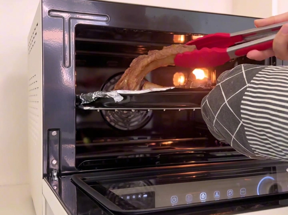 Red silicone kitchen tongs turning over pieces of baked pork ribs inside the oven tray.