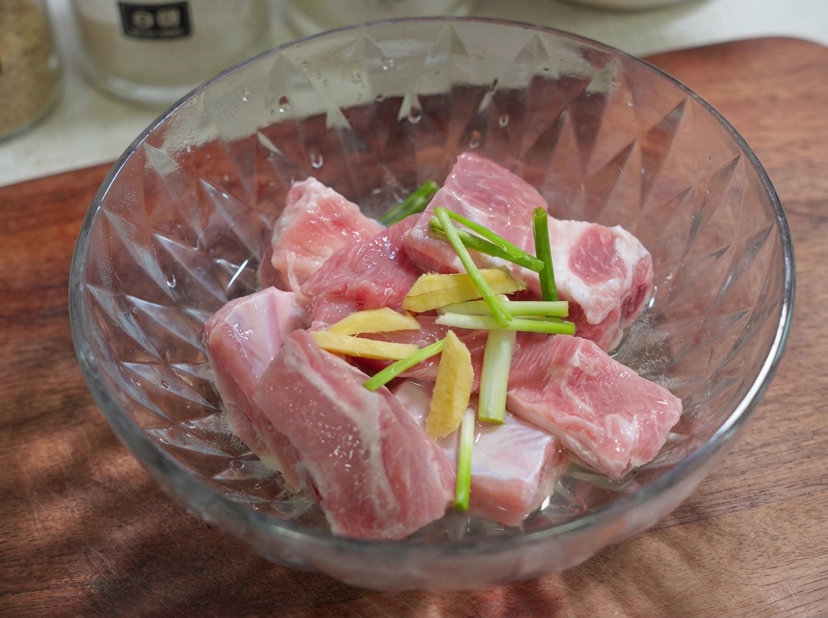 Sliced ginger and green onion pieces resting on top of raw pork ribs in a glass bowl.