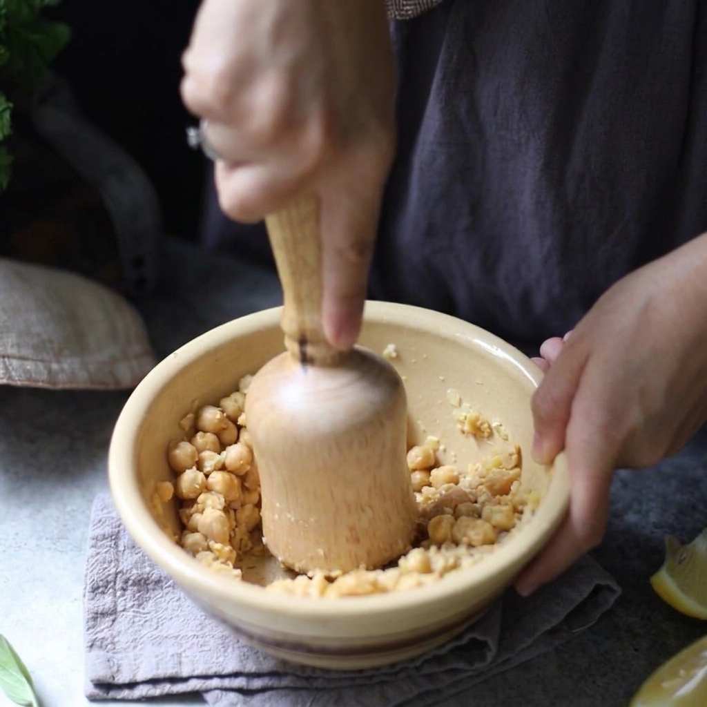 A person using a wooden pestle to manually mash boiled chickpeas inside a yellow ceramic bowl.