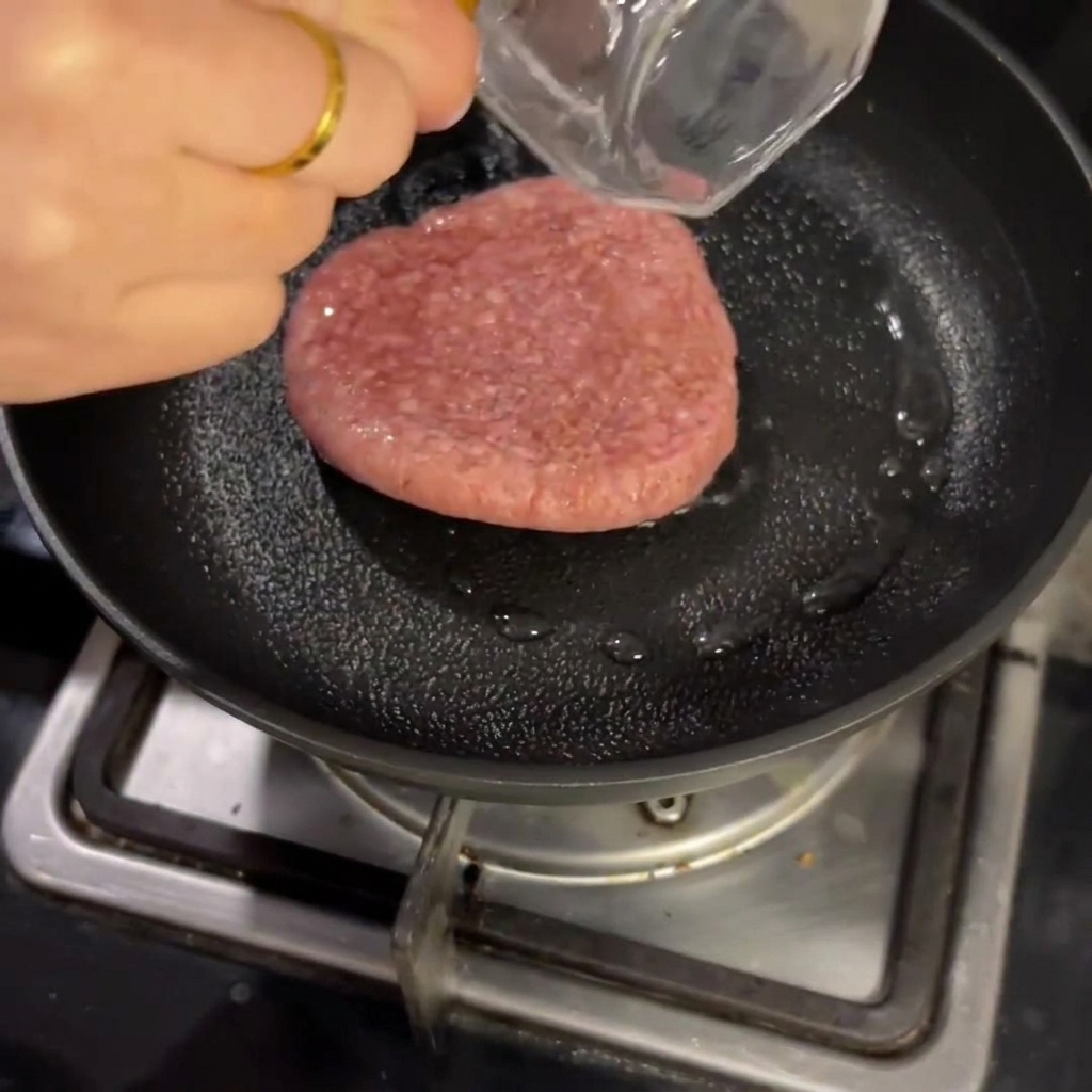 A raw beef patty cooking inside a frying pan with a small amount of liquid, covered with a lid.