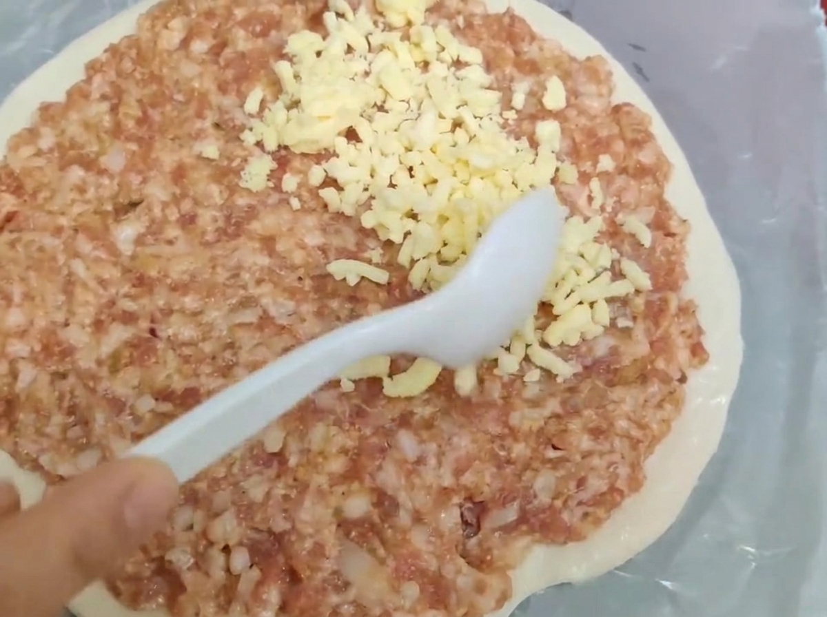 A white spoon spreading raw minced meat and shredded cheese over a round pastry dough.