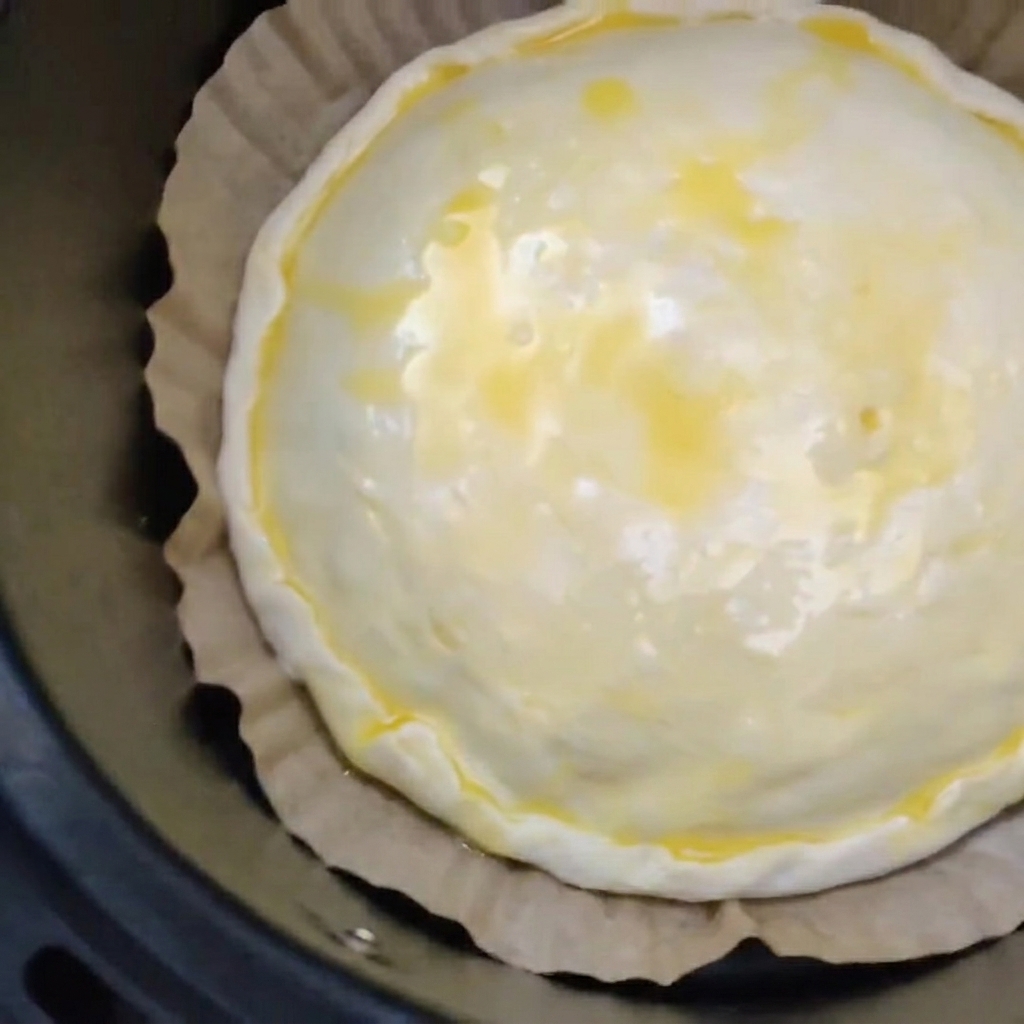 An unbaked meat and cheese pie with an egg-washed top resting on brown parchment paper inside a black air fryer basket.