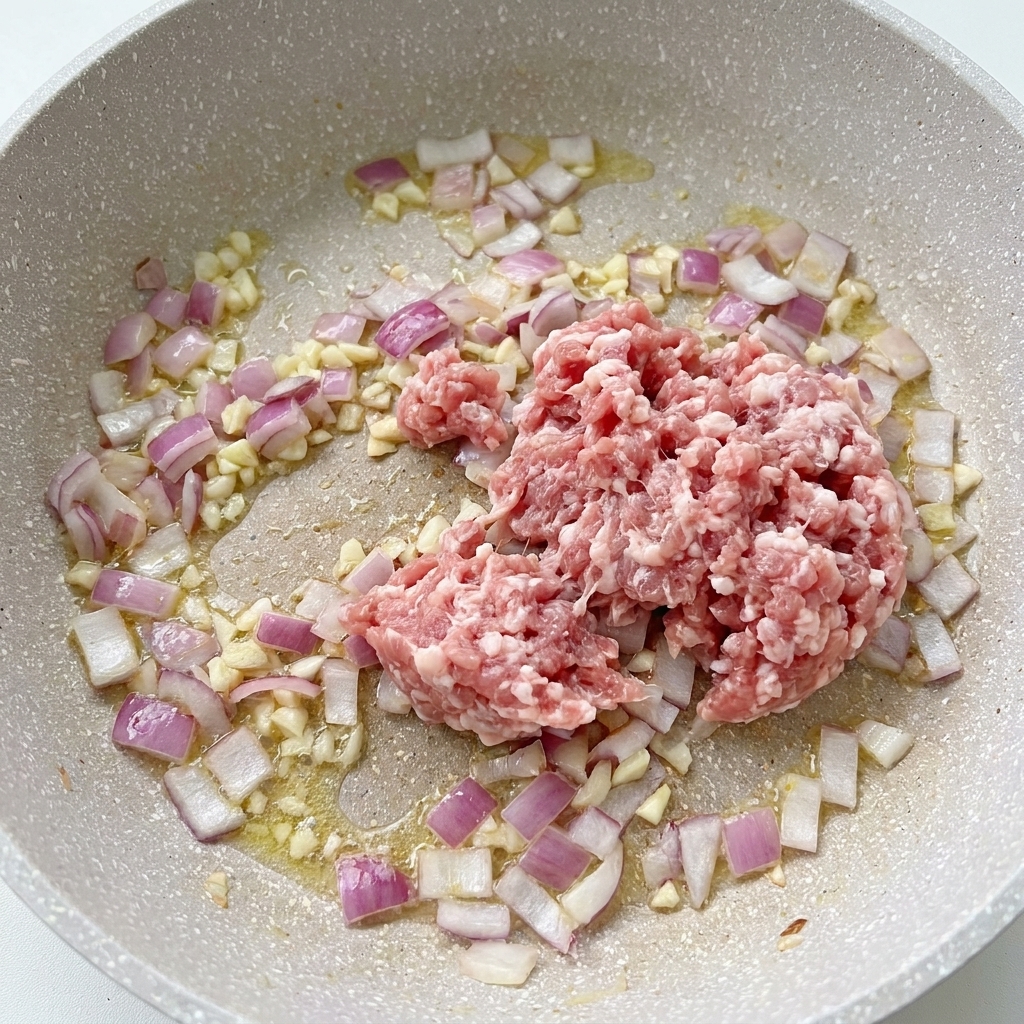 Raw minced pork sitting in a pan alongside cooking onions and minced garlic.