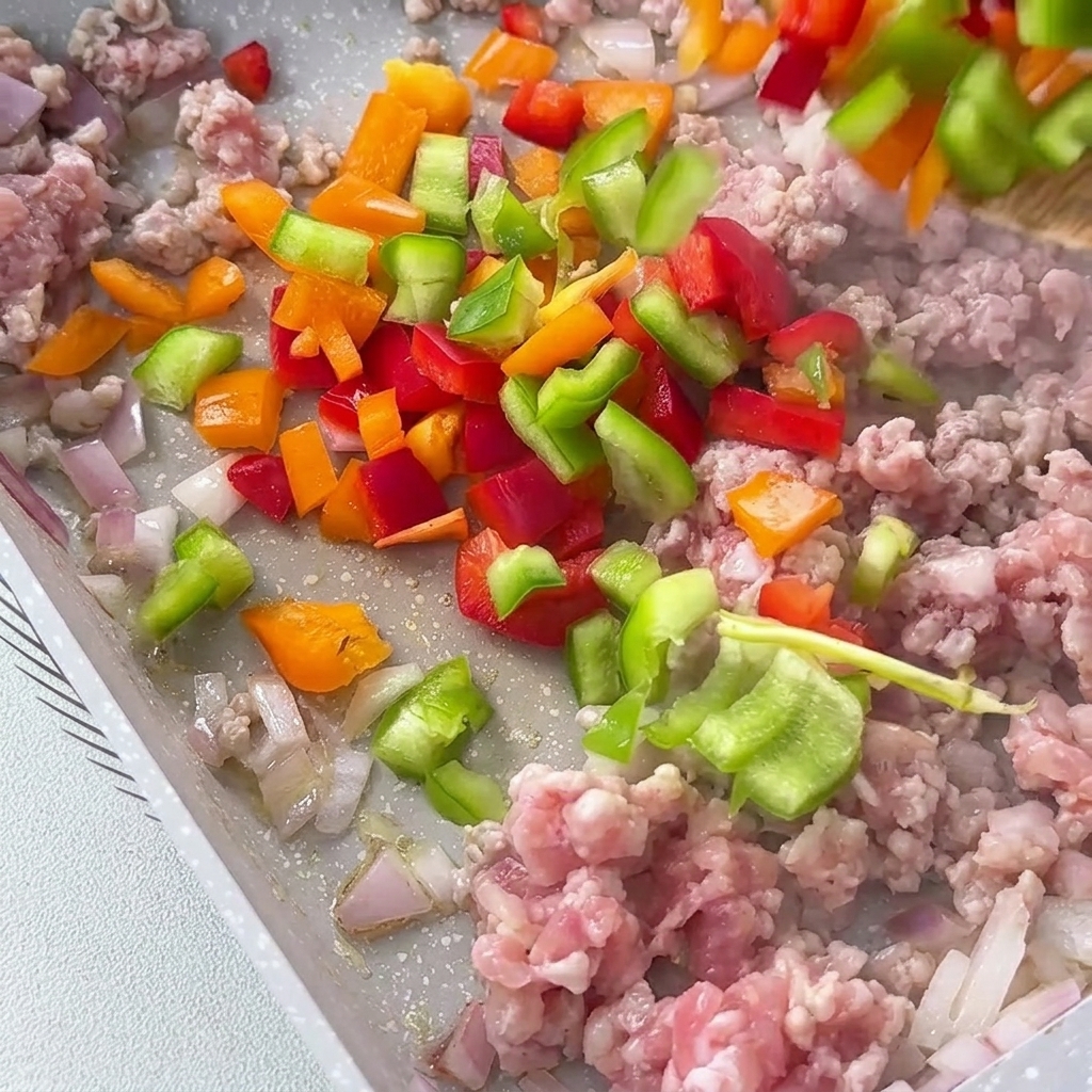 Diced red, yellow, and green bell peppers being added to the pan with the meat mixture.