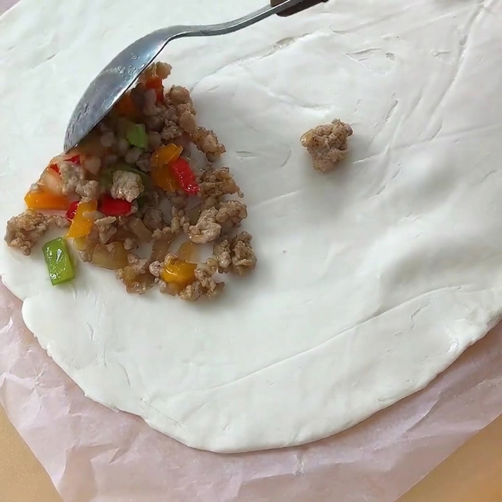 A spoonful of cooked meat filling being placed onto a circular sheet of flatbread dough on parchment paper.