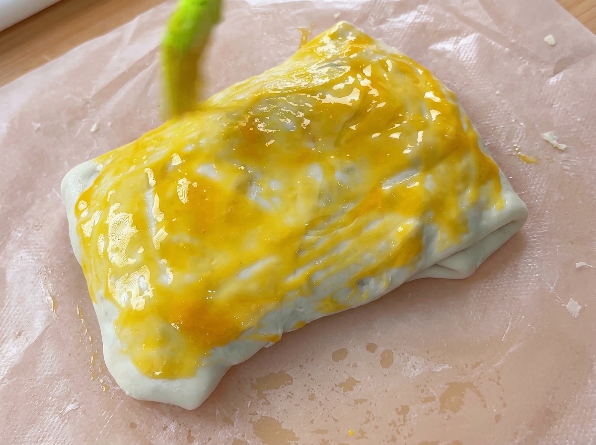A pastry brush applying a layer of bright yellow egg wash onto a folded dough parcel.