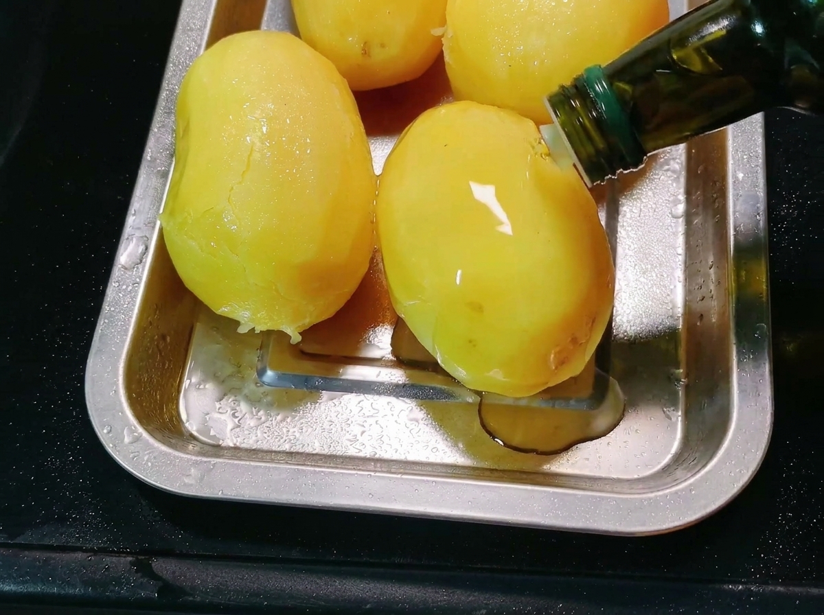Cooking oil being poured from a bottle onto peeled potatoes in a metal baking tray.