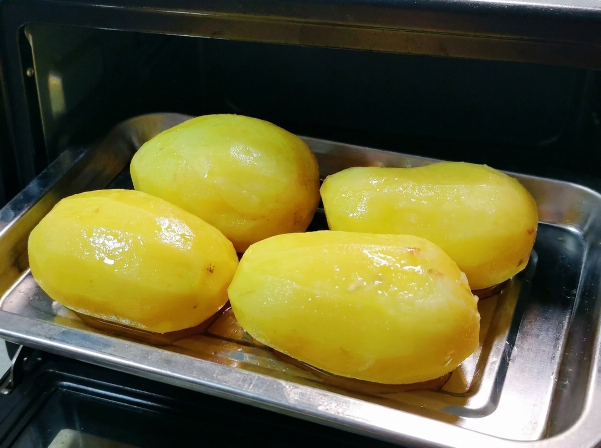A metal tray of oiled whole potatoes being placed inside a small black oven.
