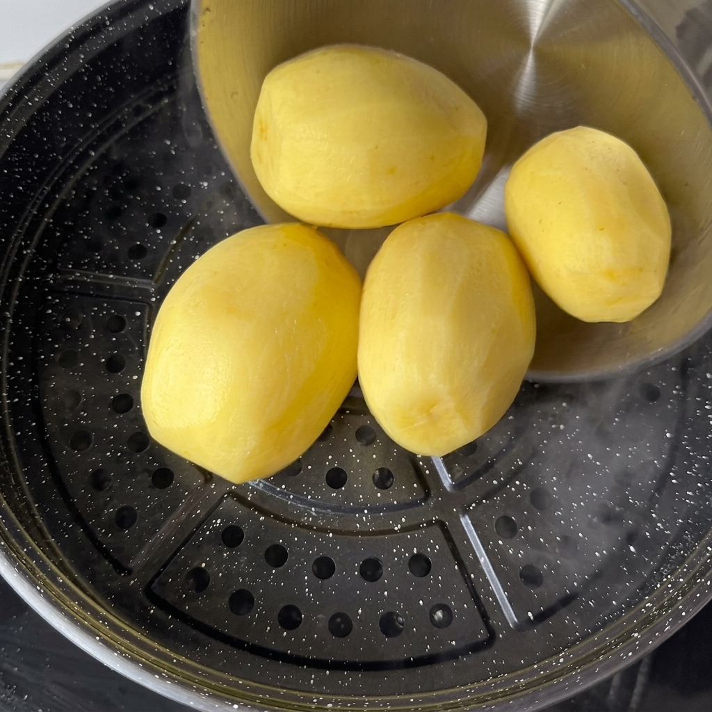 Peeled yellow potatoes being loaded onto a steaming tray over hot boiling water.