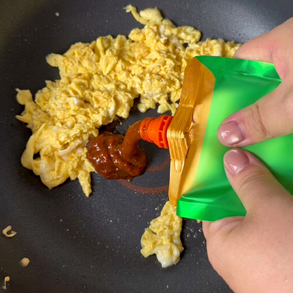 Yellow soybean paste being squeezed from a green pouch onto scrambled eggs in a pan.