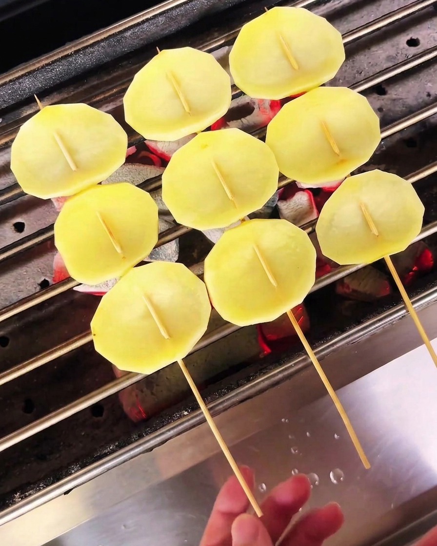 Pale potato slices on skewers being dried over a charcoal grill.