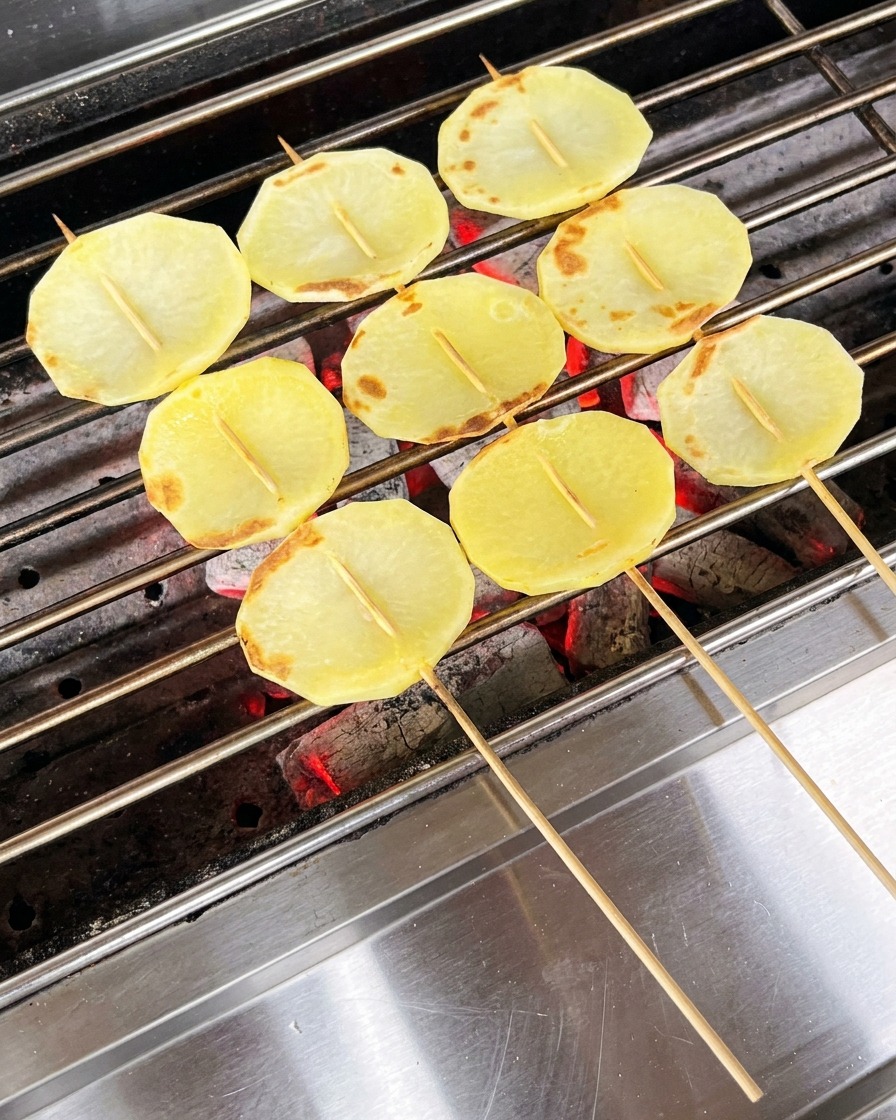 Partially cooked potato slices on the grill being coated with oil using a brush.