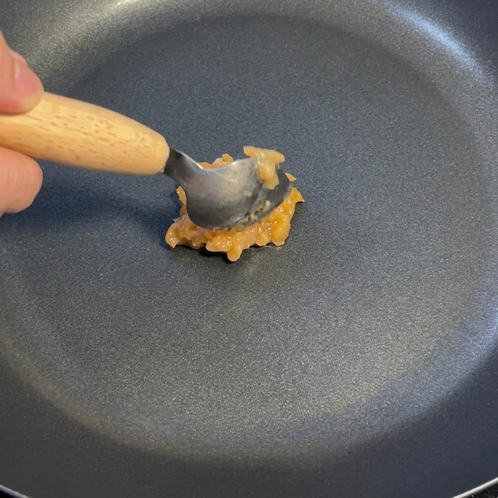 A spoonful of garlic and ginger sauce being stirred in a dark non-stick pan over low heat.