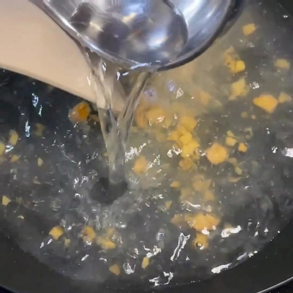 Hot water being poured from a ladle into a boiling pan containing seasoning and bits of ginger.