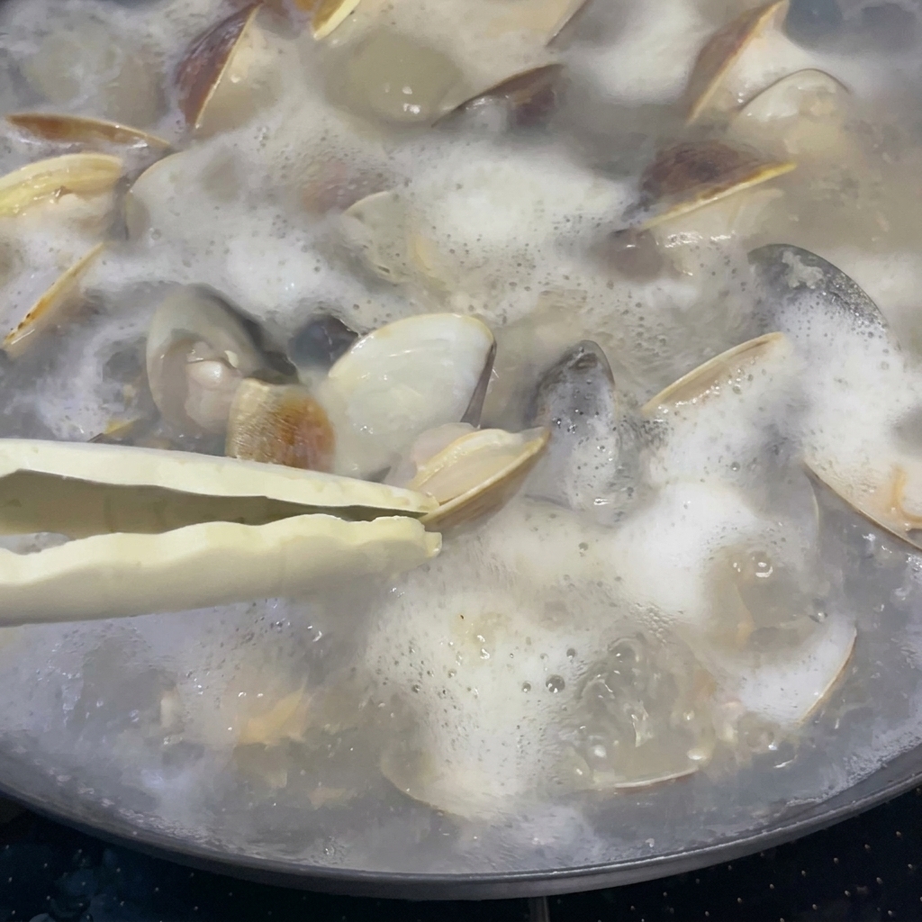 A tablespoon of rice wine being poured into a pot of bubbling clams and broth.