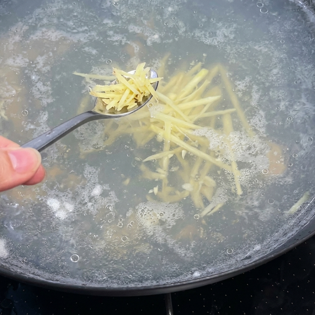 A spoonful of shredded ginger being added into a boiling pot of clear soup broth.