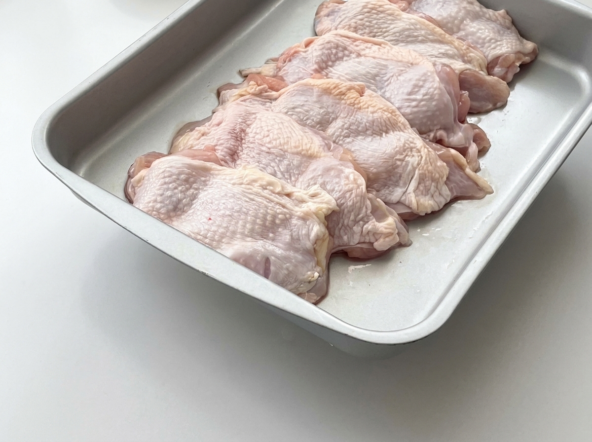 Raw chicken thighs laid out in a metal tray with fresh ginger slices placed on top.