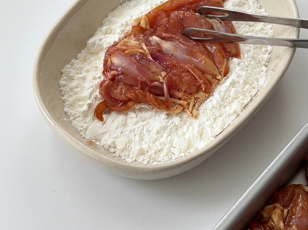 A piece of marinated chicken being handled with tongs in a bowl filled with white cornstarch.