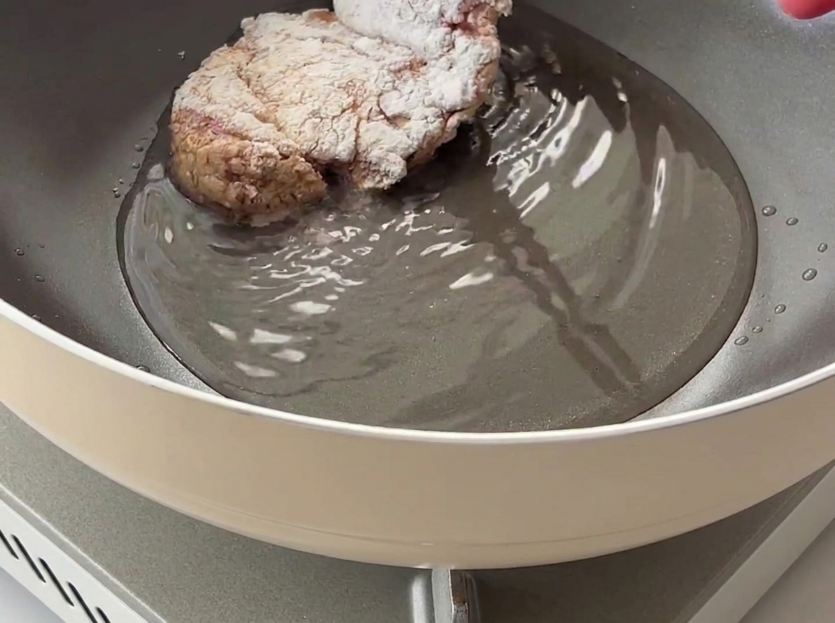 A cornstarch-coated chicken thigh being placed into a pan with hot cooking oil.