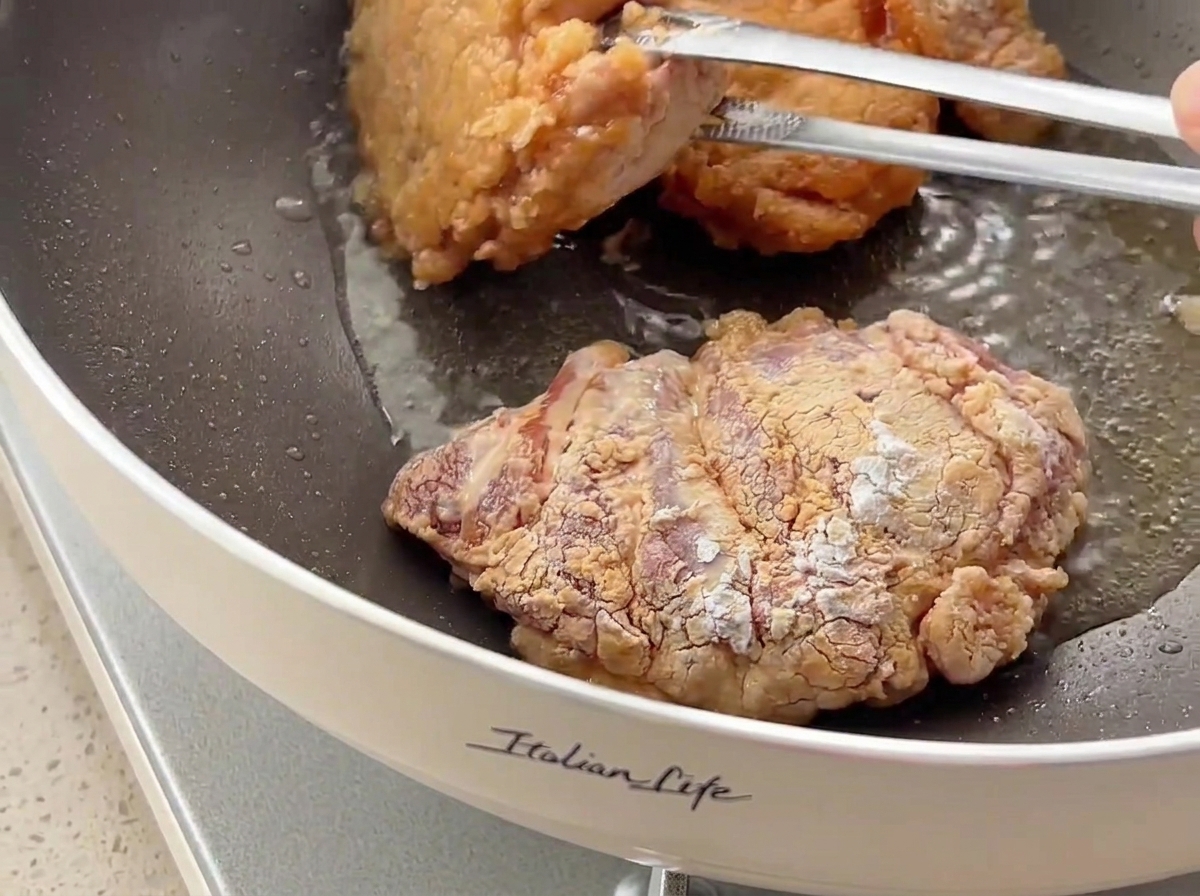 Chicken pieces being flipped with tongs in a frying pan, showing a golden-brown crispy crust.