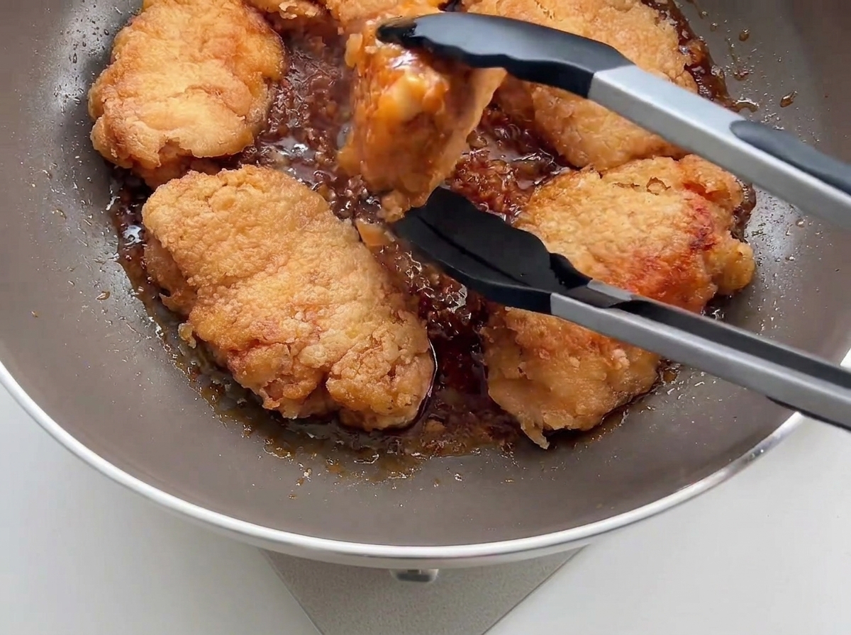 Crispy fried chicken being placed into a pan with garlic sauce using black kitchen tongs.