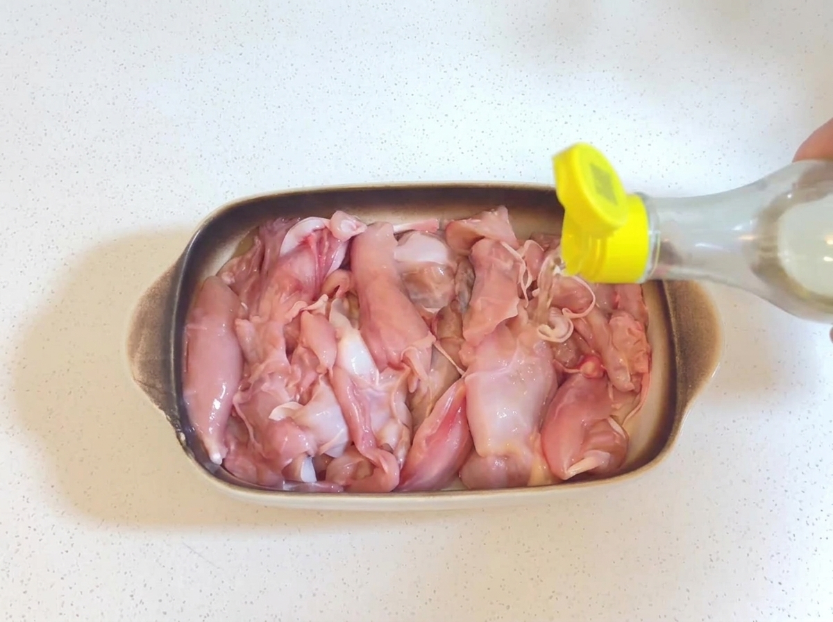 Clear cooking wine being poured from a small bottle with a yellow cap onto raw chicken pieces in a baking dish.