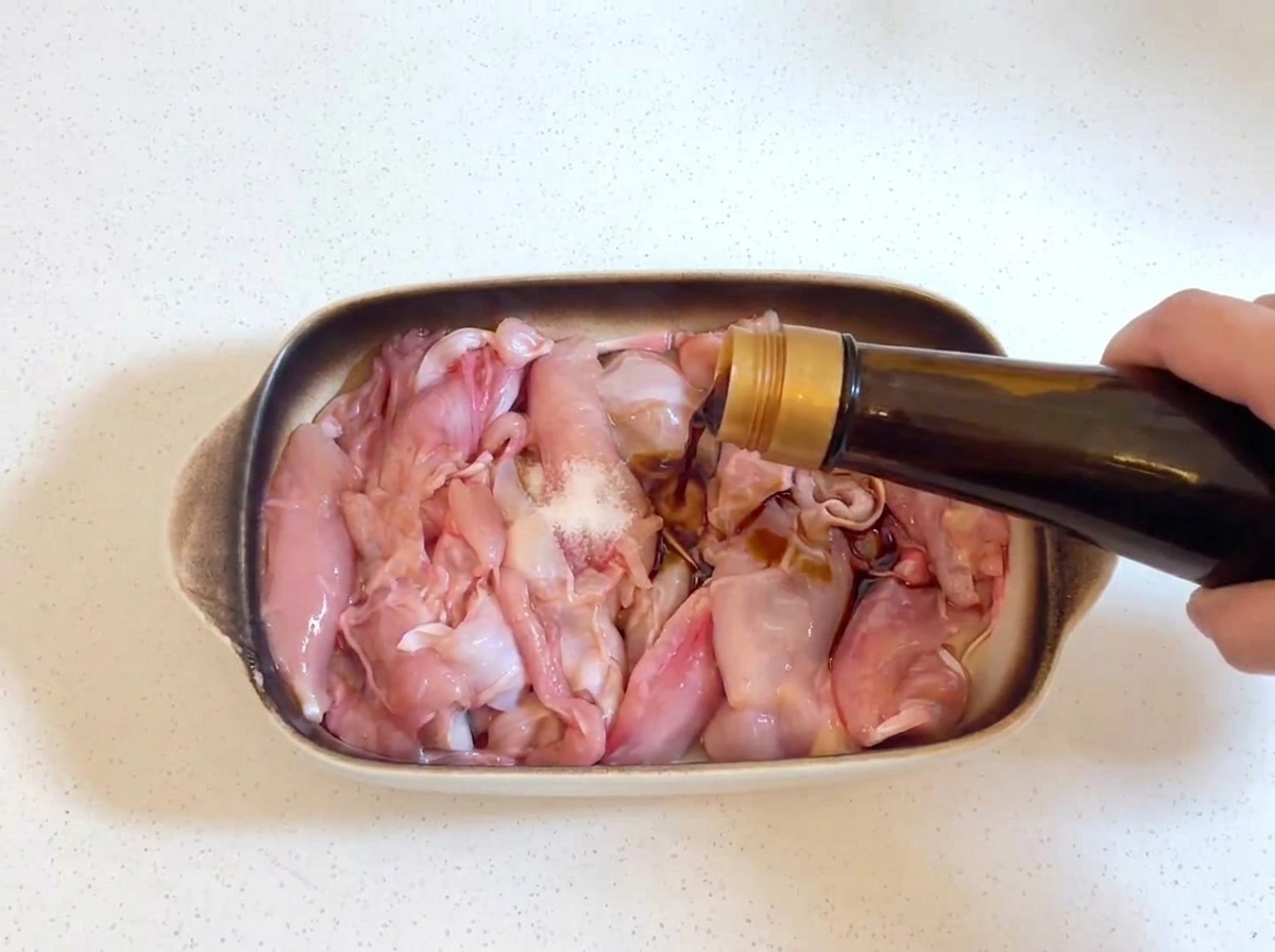Dark soy sauce being poured from a glass bottle onto raw chicken pieces, with a small pinch of salt visible on the chicken.