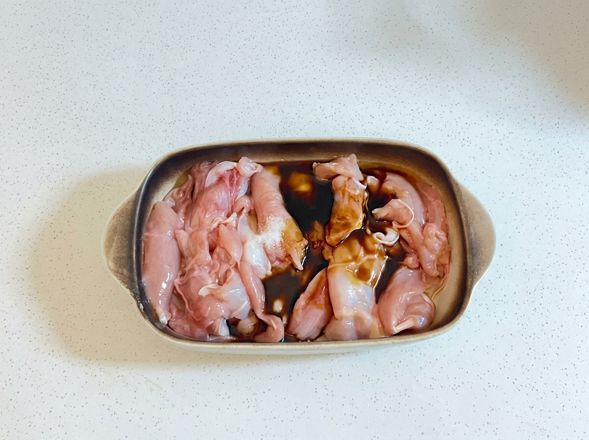 Dark cooking liquids and oil being poured over raw chicken pieces in a baking dish.