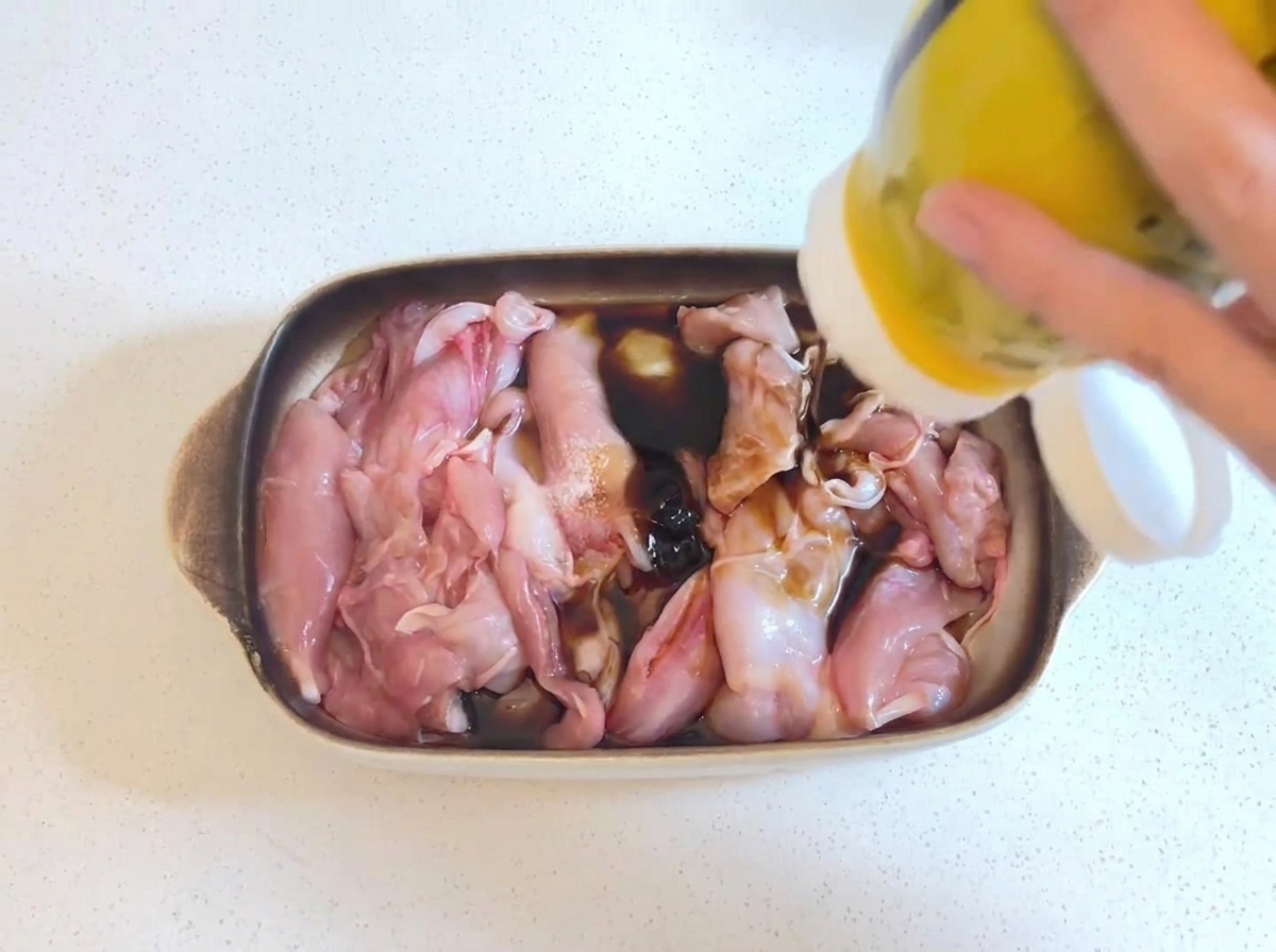 Thick dark oyster sauce being squeezed from a yellow bottle onto raw chicken pieces sitting in a soy sauce marinade.