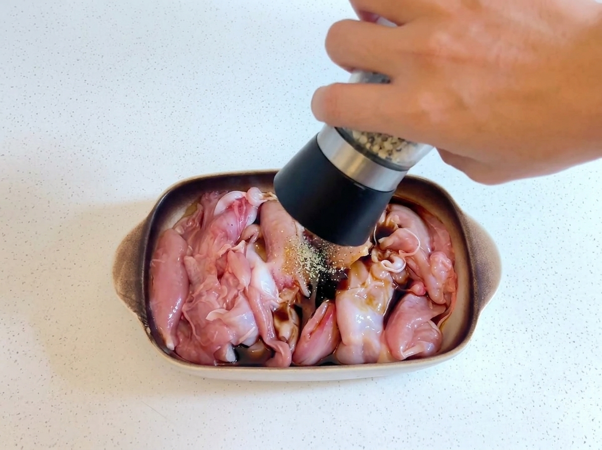 A hand holding a pepper grinder and grinding fresh pepper over raw chicken marinating in a dish.