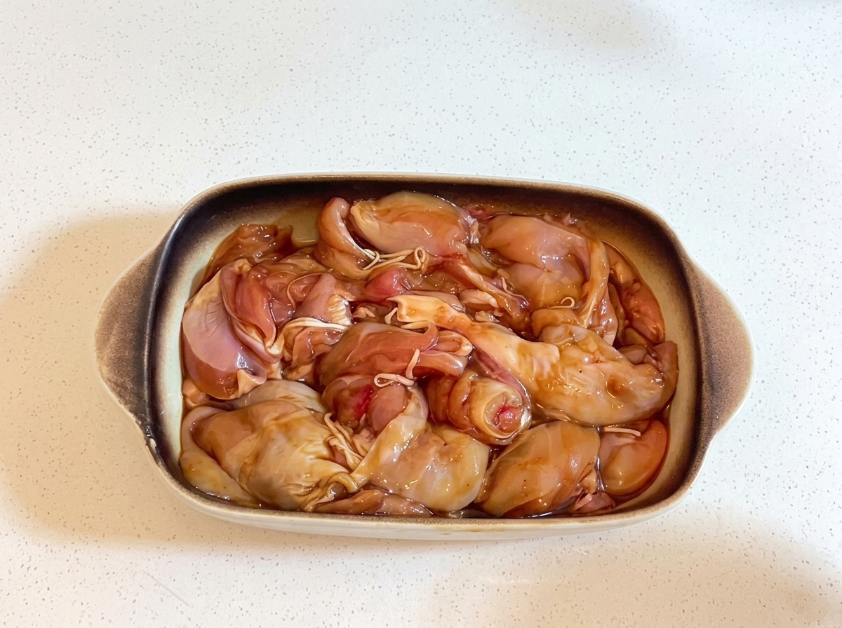 A rectangular ceramic baking dish filled with marinated chicken pieces, tightly covered with transparent plastic wrap.