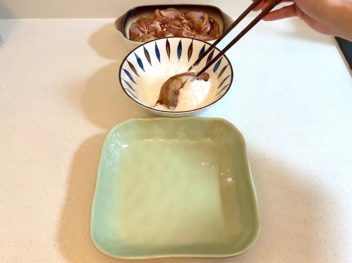 Chopsticks holding a piece of marinated chicken inside a bowl filled with white starch.