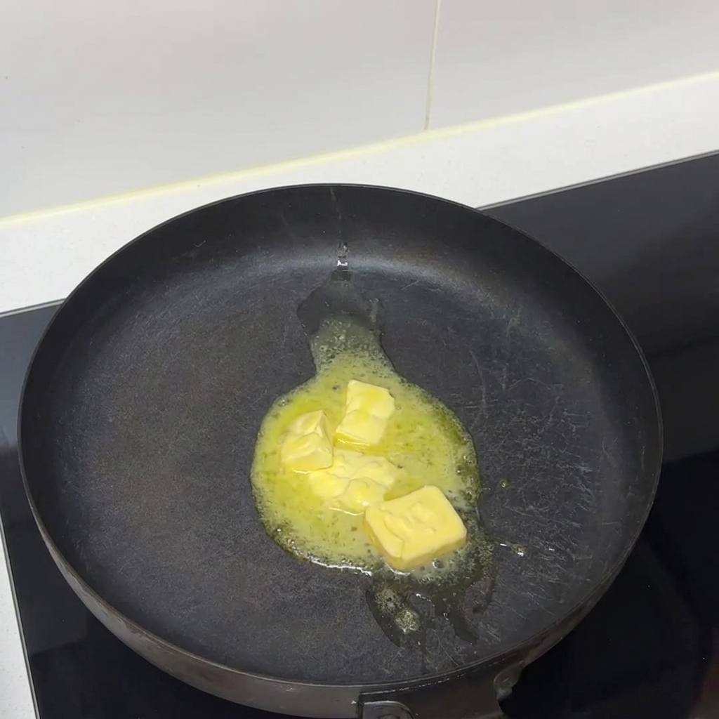 Butter pieces melting into liquid in a dark frying pan on a stovetop.
