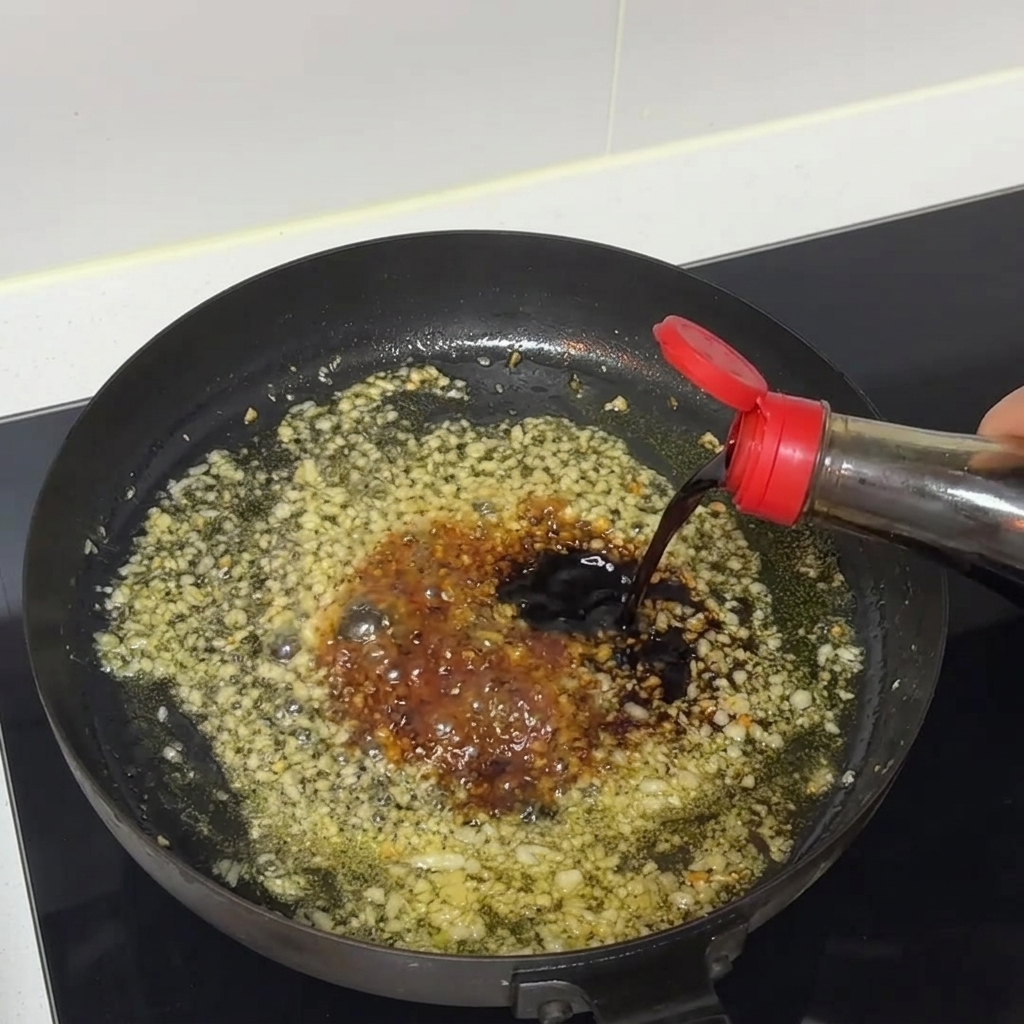 Dark soy sauce pouring from a bottle into a sizzling pan of minced garlic and sauce.