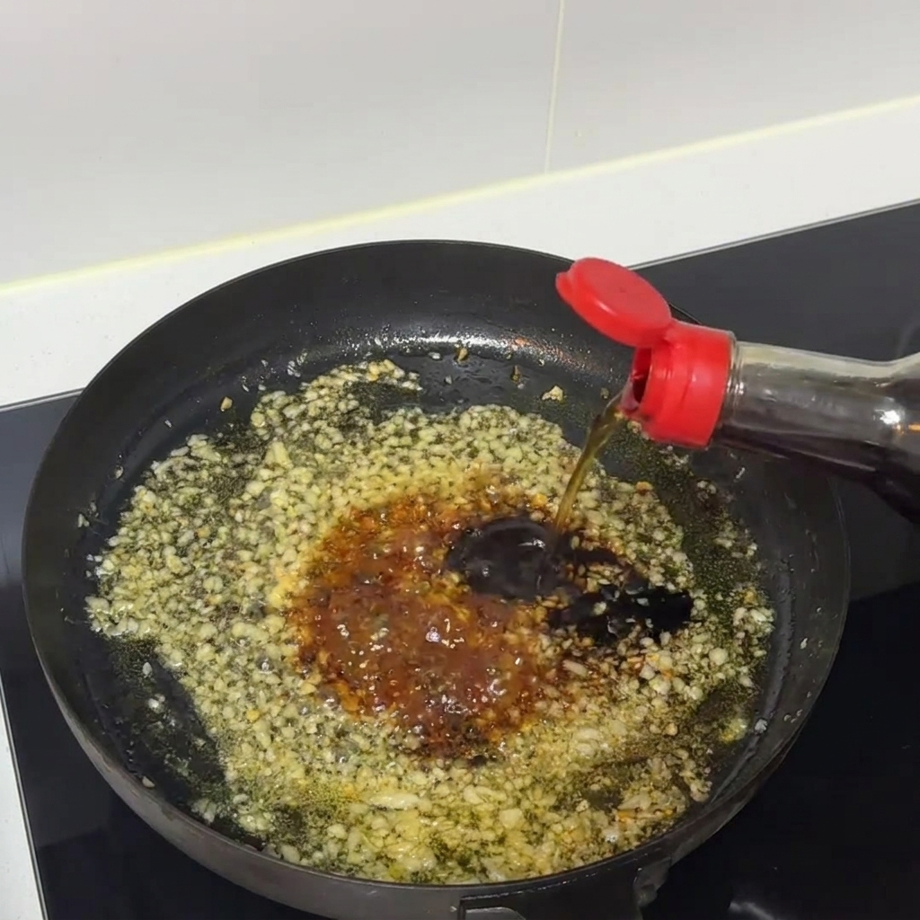 Dark sauce mixture simmering and being stirred inside a frying pan with minced garlic.