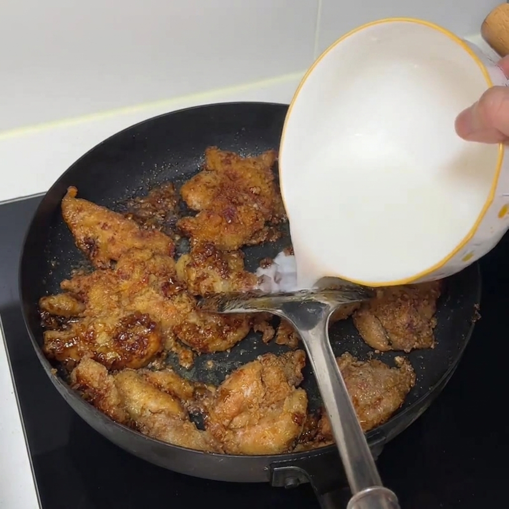 Starch water being poured from a white bowl into a frying pan filled with cooked chicken pieces.