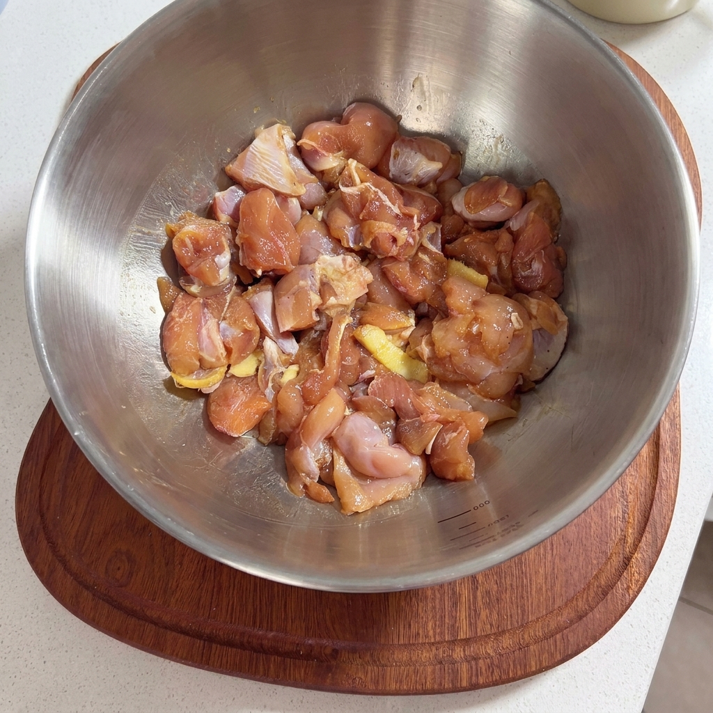 A metal bowl filled with raw chicken thigh pieces seasoned with marinade sauce and ginger slices.