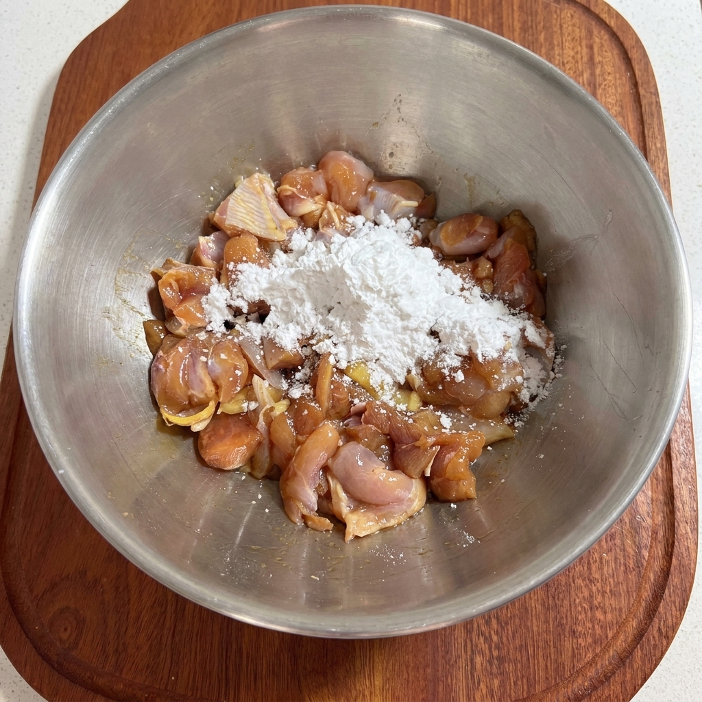 A hand mixing white starch into seasoned chicken pieces inside a metal bowl.