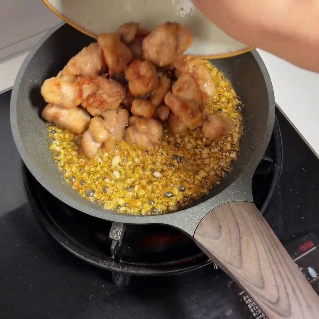 Pre-fried chicken pieces being poured from a plate into a frying pan filled with garlic sauce.