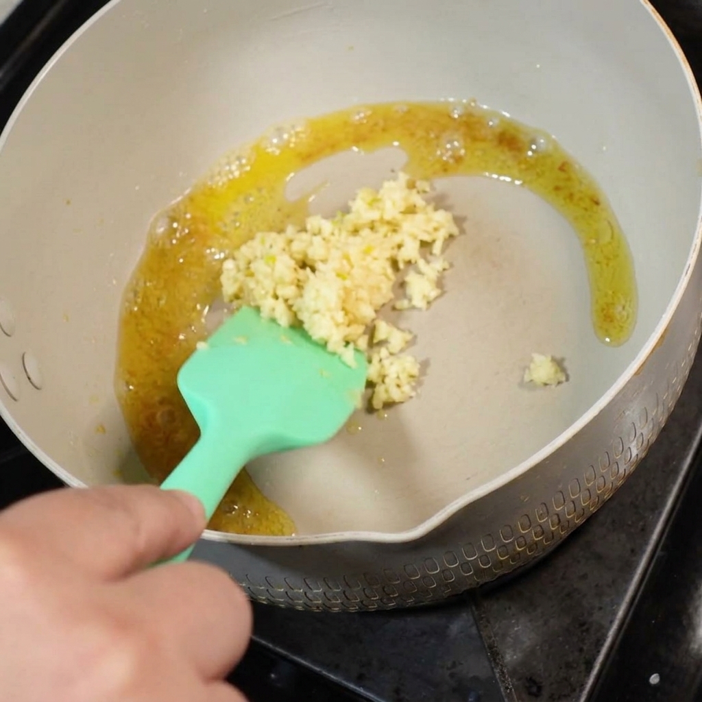 Fresh minced garlic being added to melted butter in a pan, stirred with a green silicone spatula.