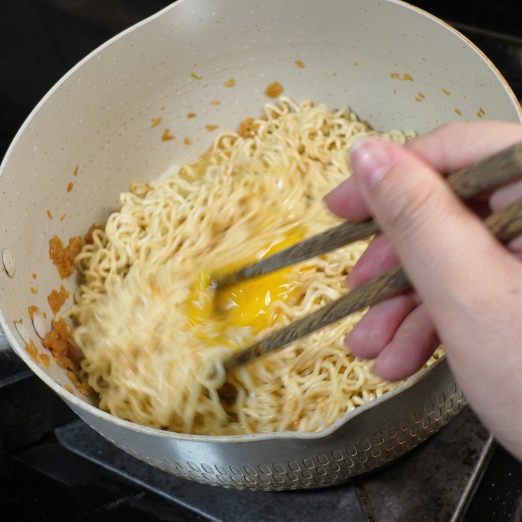 An egg being stirred and scrambled into noodles inside a pan using wooden chopsticks.