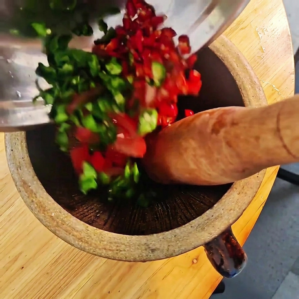 Chopped green and red chilies and tomatoes being crushed in a wooden mortar with a pestle.