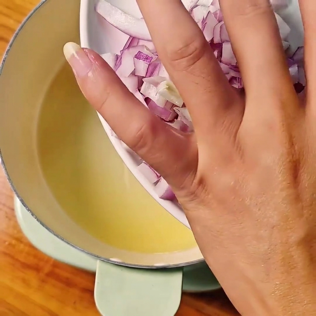 Diced red onions being poured from a white plate into a pot of heated oil.