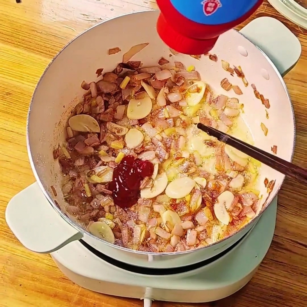 Red tomato paste being squeezed out of a bottle into a pot of sautéed onions and garlic.