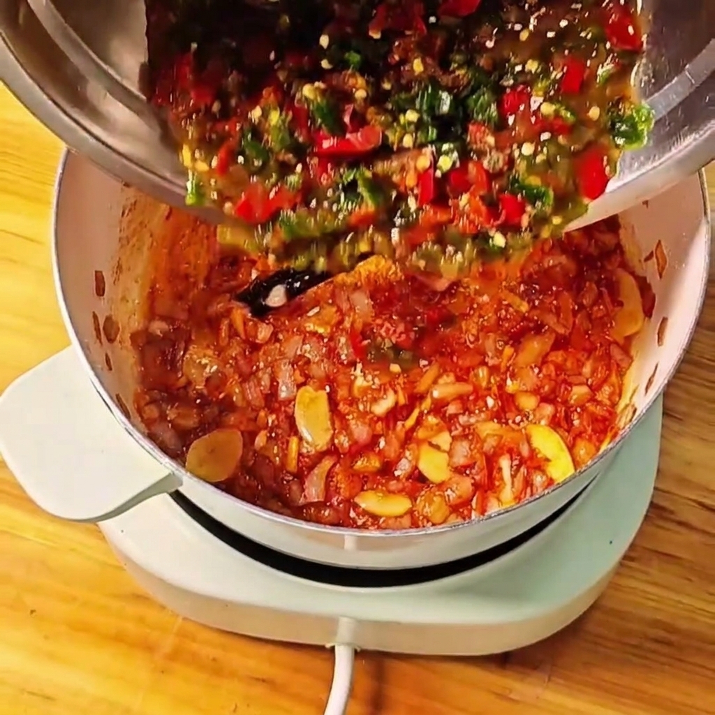 Mashed tomatoes and green and red chilies being poured from a metal bowl into the cooking pot.