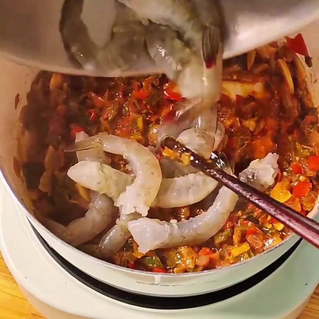 Raw grey whole shrimp being dropped into a simmering red tomato sauce in a white pot.