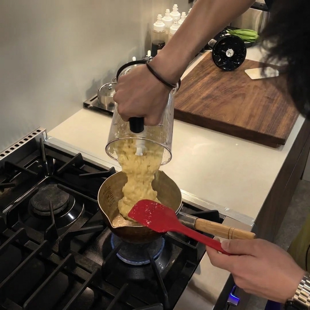 Pouring a smooth, pale yellow blended puree from a blender pitcher into a small metallic saucepan on a gas stovetop.
