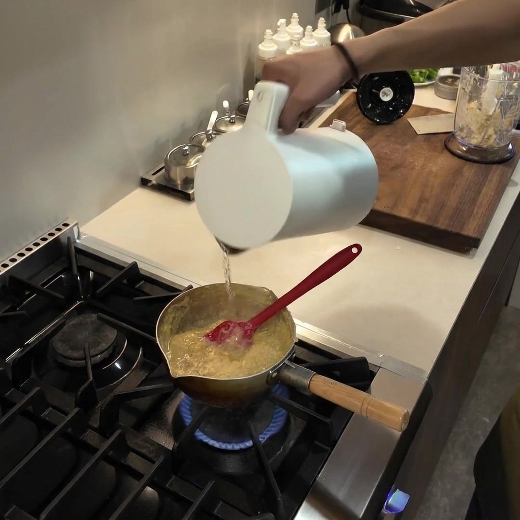 Pouring water from a white pitcher into a saucepan containing a thickening, simmering fufu mixture.