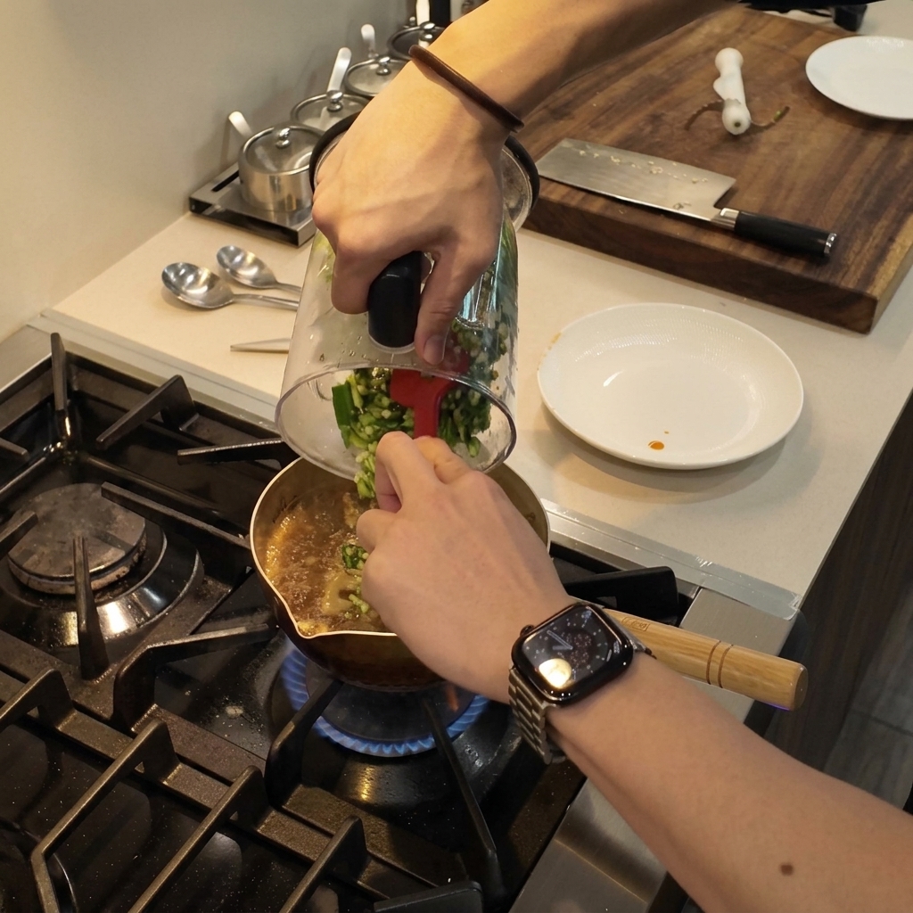 Minced green okra being scraped from a blender cup into a pot of simmering stew using a red spatula.