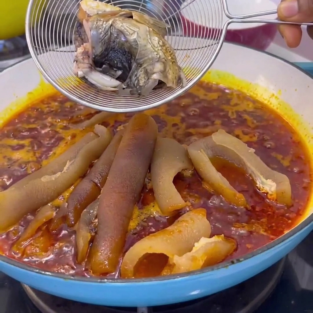 A wire strainer holding cooked fish pieces lowered into a pot with simmering red sauce and pork skin.